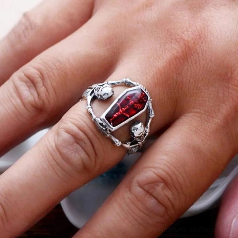 Steampunk coffin skull ring worn on human finger with small skull accents on band, vintage silver and red enamel finish - eye-level side view on dark brown background
