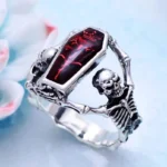 Steampunk coffin skull ring with red enamel inlay and skeleton frames, vintage silver tone, displayed on light blue desktop with flower decor - oblique overhead view