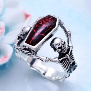 Steampunk coffin skull ring with red enamel inlay and skeleton frames, vintage silver tone, displayed on light blue desktop with flower decor - oblique overhead view