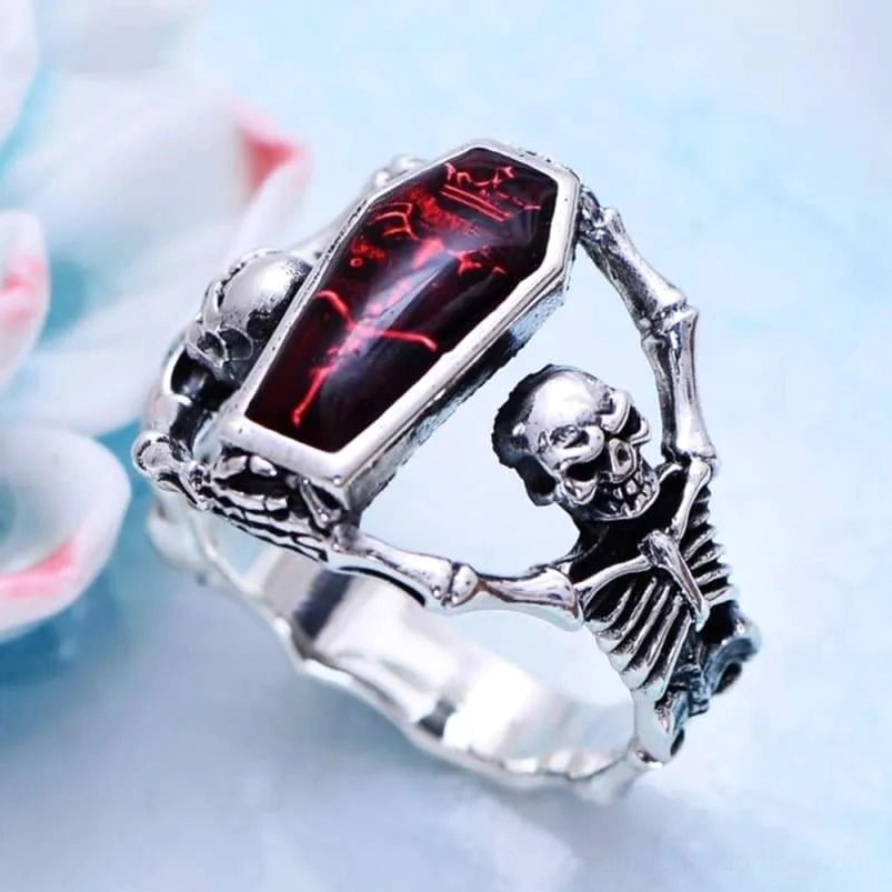 Steampunk coffin skull ring with red enamel inlay and skeleton frames, vintage silver tone, displayed on light blue desktop with flower decor - oblique overhead view