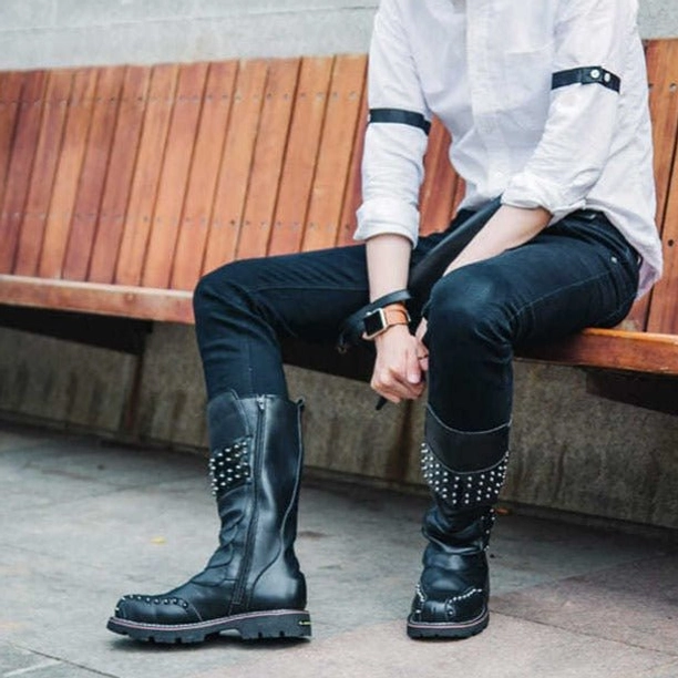 Man wearing black steampunk studded mid-calf combat boots outdoors on wooden bench, street styling demonstration, casual daily wear with rolled cuffed jeans, showcasing wearable versatility
