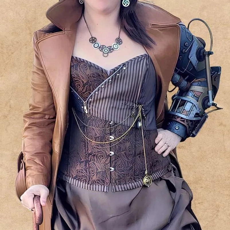 Female model wearing steampunk corset outfit with brown trench coat, decorative chains, and paisley corset details, styled for cosplay and conventions