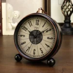 Steampunk desk clock with black metal frame, bronze accents, Gothic numerals on aged dial, and spherical metal feet—front left perspective on wooden study desk