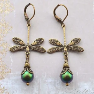 Pair of matching steampunk dragonfly earrings featuring engraved bronze metal with detailed wing carvings and green iridescent pearl drops on light pink vintage textured background with gold glitter accents
