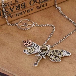 Steampunk dragonfly necklace pendant with gear detailing displayed on vintage wooden table with antique book, showing mixed metal finish and intricate mechanical wing design