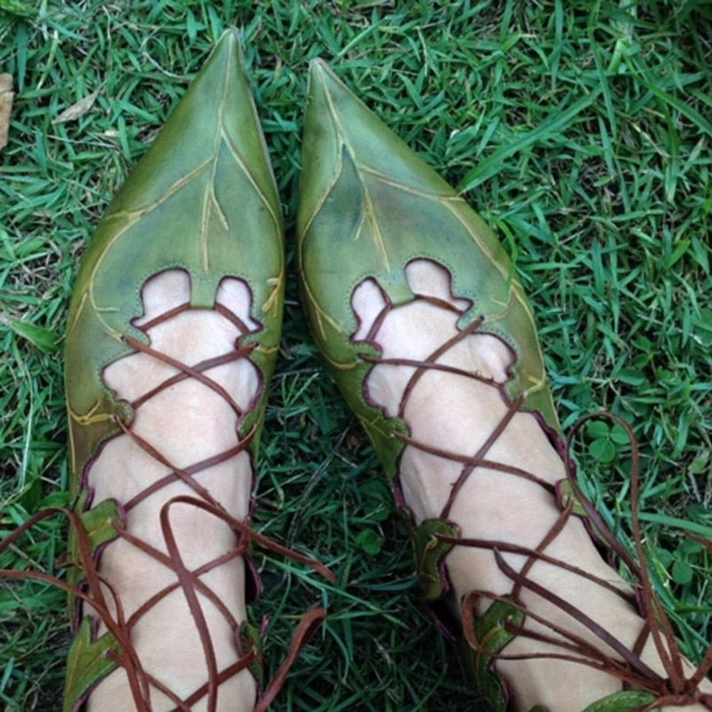 Pair of green pointed elf lace-up flats with intricate brown crisscross lacing pattern, overhead view on grass