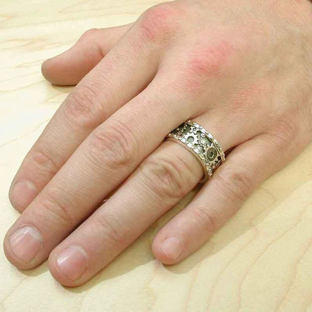 Steampunk carved gear ring worn on adult male middle finger in lifestyle shot, displaying worn silver gear band and authentic wearability on light wood tabletop background