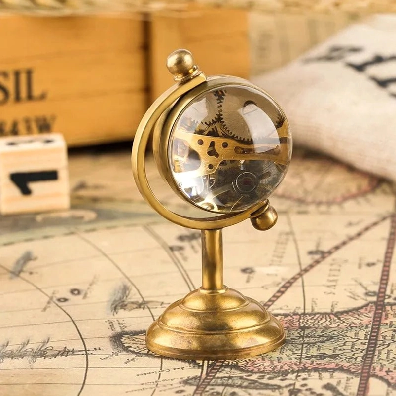 Steampunk globe ornament clock with clear glass sphere, visible internal brass mechanical movement, brass metal frame, three-quarter view on vintage map surface