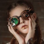 Green lens steampunk goggles with spiked metal frame and side magnifier worn by model with braided hair
