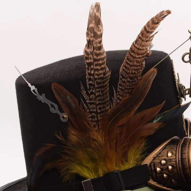 Close-up detail of steampunk hat featuring pheasant feathers, clock hand ornament, bronze goggle strap, and green-trimmed black ribbon band