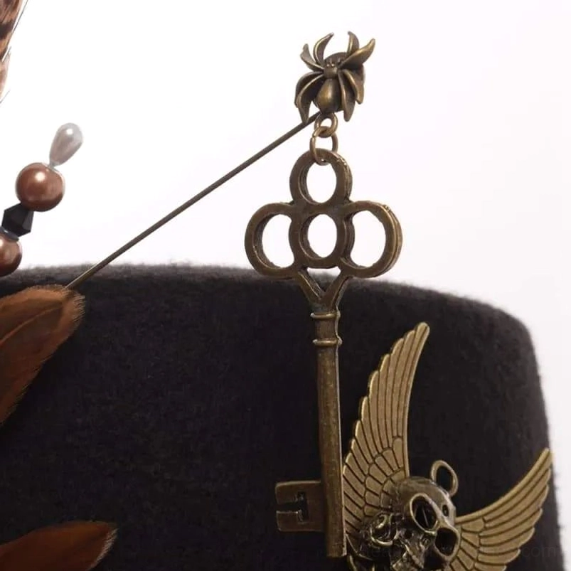 Steampunk hat decoration side close-up showing winged skull key ornament, beaded metal pin accent, and brown pheasant feather detail