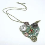 Steampunk heart necklace with mixed metal gears, patina green frame, red central gemstone, and wing ornaments displayed in oblique side view on white background