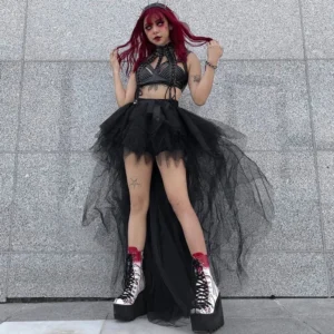 Black gothic high-low tulle skirt with dramatic back train, paired with lace-up bustier and platform boots, full-body front view on grey stone wall background