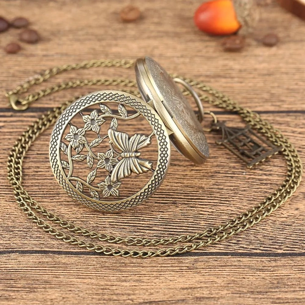 Flip-style steampunk hollow pocket watch showing carved flower and butterfly cover with birdcage pendant charm and coiled metal chain, side-top view