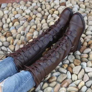Dark brown steampunk knee-high boots with lace-up front and side zipper, featuring buckle straps and block heel, worn with blue jeans on pebble stone ground