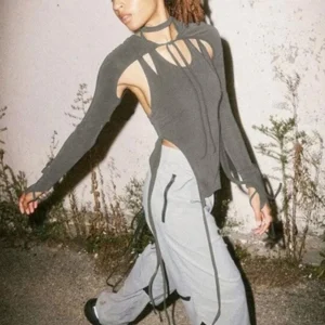 Steampunk layered long-sleeve cutout top with split side drawstrings and halter strap details in dark grey knit, worn by Black female model with dreadlocks in three-quarter outdoor walking view against textured concrete wall