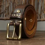 Full-grain dark brown leather steampunk belt with antique brass double pin buckle, cross-stitch detailing, and engraved strap patterns, displayed on wooden surface—front perspective view showing industrial craftsmanship and vintage aesthetic