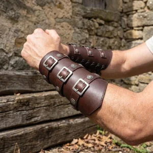 Brown steampunk leather forearm bracers featuring silver square buckles and rivet trim details, showcased on male model in outdoor rustic setting with natural stone and wood textures emphasizing industrial aesthetic