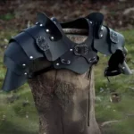 Black steampunk leather shoulder and chest armor featuring silver rivets and ornate carved buckles, displayed on wooden tree stump in lush woodland setting, left three-quarter angle detail view highlighting layered construction and metallic hardware accents