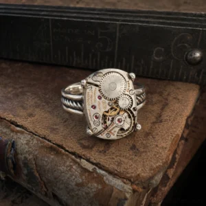 Steampunk mechanical gear ring with silver twisted cable band and watch movement details, displayed on vintage book with metal ruler, showing intricate gears and red gemstone accent