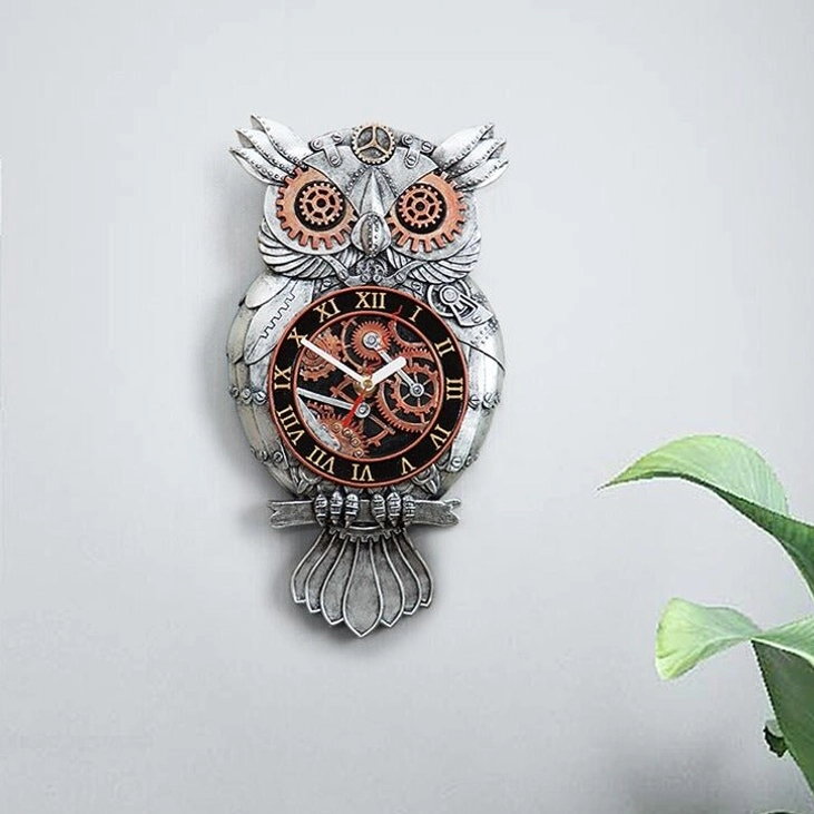 Compact steampunk owl wall clock full view against light gray wall with single plant leaf, demonstrating overall proportions and vintage-inspired design aesthetic