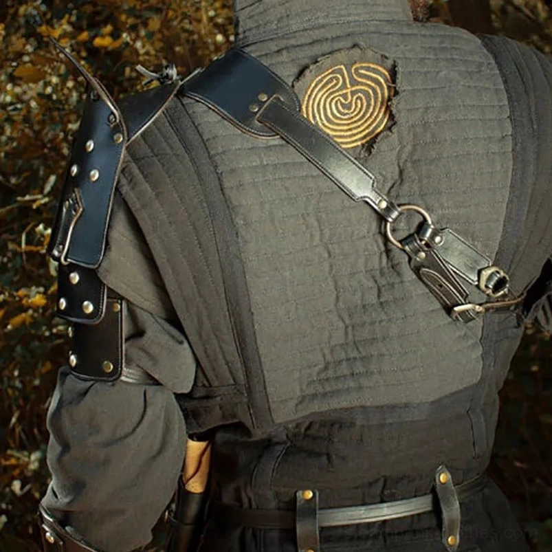 Back view of steampunk shoulder armor displaying cross-body strap fastener system and decorative golden spiral embroidery pattern on gray linen coat, outdoor woodland backdrop