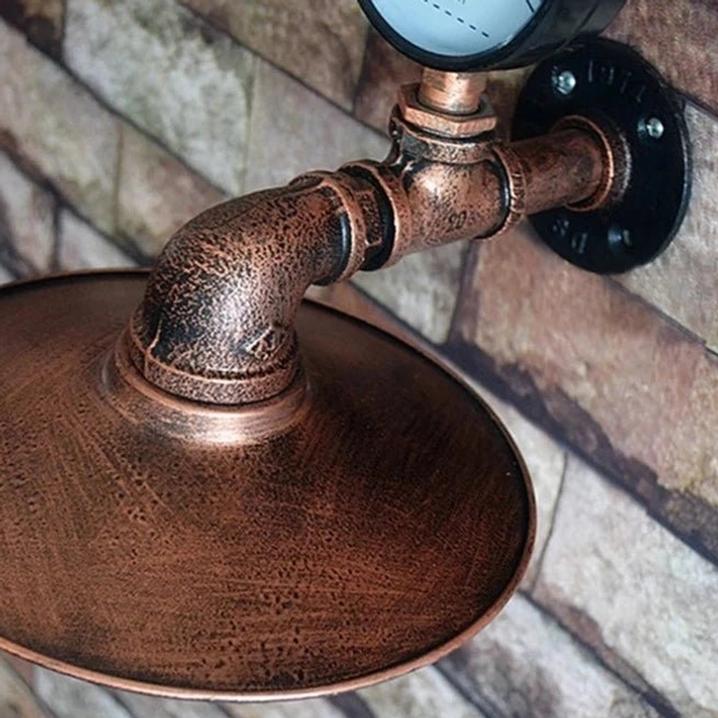 Industrial pipe wall lamp hardware mounted on brick wall, showing distressed copper pipe joints, black wall mount plate and conical metal shade, side three-quarter view