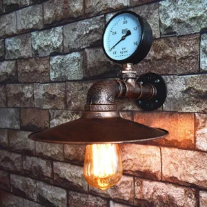 Steampunk industrial pipe wall sconce with pressure gauge mounted on brick wall, showing distressed copper finish and exposed Edison filament bulb, front three-quarter view