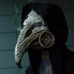 Steampunk plague doctor mask worn by male model in black hooded robe, left side close-up showcasing textured bone-like beak and ornate metal gas filter with atmospheric smoke effect