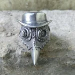 Steampunk plague doctor ring frontal view showing antique silver finish with bowler hat crown, dual mechanical goggles, stitched mask details and pointed beak design on stone background