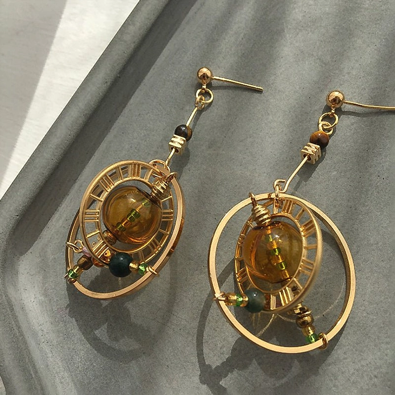 Steampunk planet earrings flat lay on gray ceramic tray featuring Roman numeral hollow frame, amber center bead, and multi-color accent beads with gold ear wires