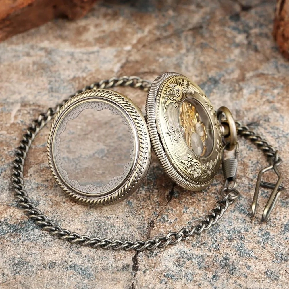 Steampunk pocket watch case cover and detached body showing bronze carved metal, clear crystal display window with floral engravings, and linked metal chain from overhead angle