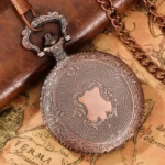 Vintage steampunk pocket watch with embossed copper-red case displayed on antique map and leather notebook, showing floral frame and shield-shaped central engraving from top-down left perspective