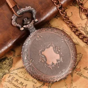 Vintage steampunk pocket watch with embossed copper-red case displayed on antique map and leather notebook, showing floral frame and shield-shaped central engraving from top-down left perspective