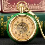 Green steampunk pocket watch with open heart mechanical movement and gold hardware, shown front view against vintage books and leather surface