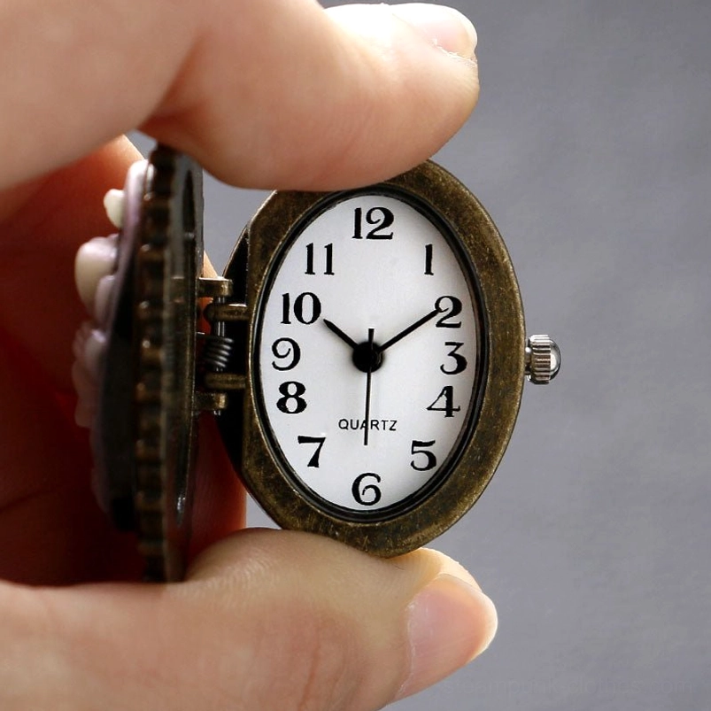 Functional quartz analog clock face inside pocket watch locket pendant, held in human hand against plain gray background, showing working timepiece mechanism