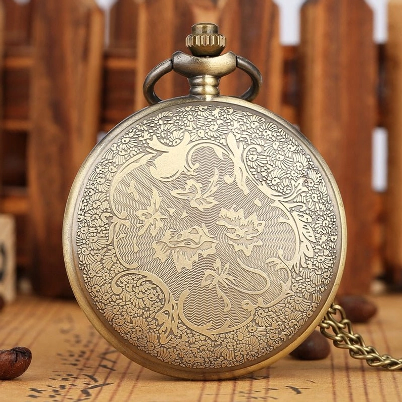 Closed steampunk pocket watch rear view showing bronze tone with full floral engraved back cover and attached metal chain against wooden fence background