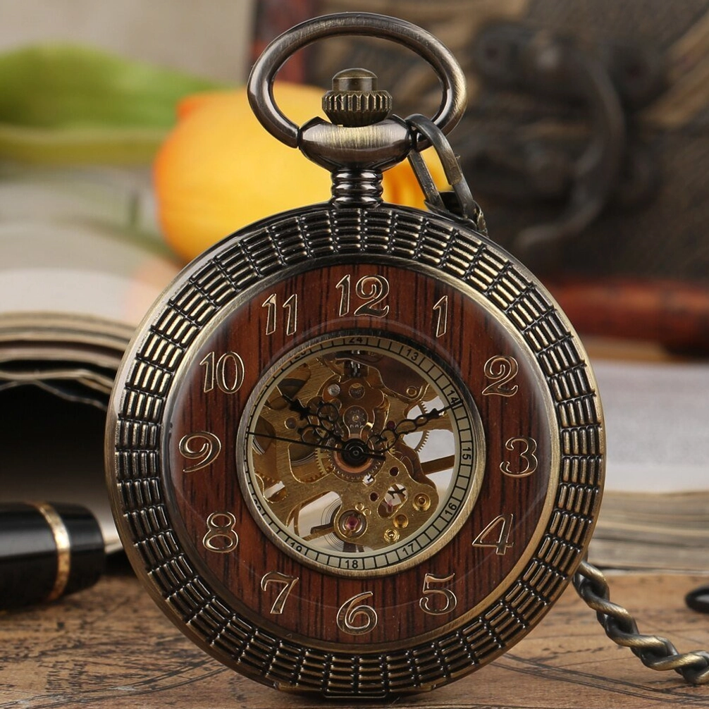 Steampunk pocket watch with exposed skeleton movement and wooden dial inlay, displayed on vintage desk with open book and yellow flower, showing front close-up of bronze metal frame