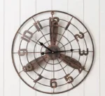 Steampunk propeller wall clock with distressed bronze finish mounted on white shiplap wall, showing full frontal view with cut-out Arabic numerals and industrial radial grid frame