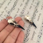 Close-up side view of steampunk raven skull dangle earrings with silver hooks and weathered off-white cracked texture on vintage musical score background