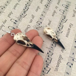 Close-up side view of steampunk raven skull dangle earrings with silver hooks and weathered off-white cracked texture on vintage musical score background