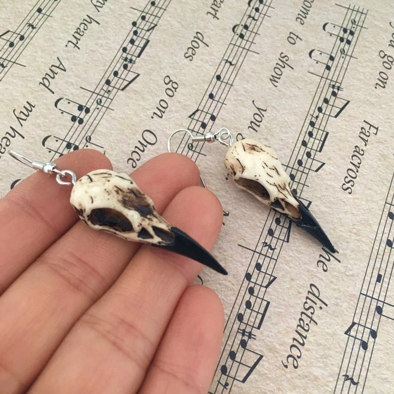 Close-up side view of steampunk raven skull dangle earrings with silver hooks and weathered off-white cracked texture on vintage musical score background
