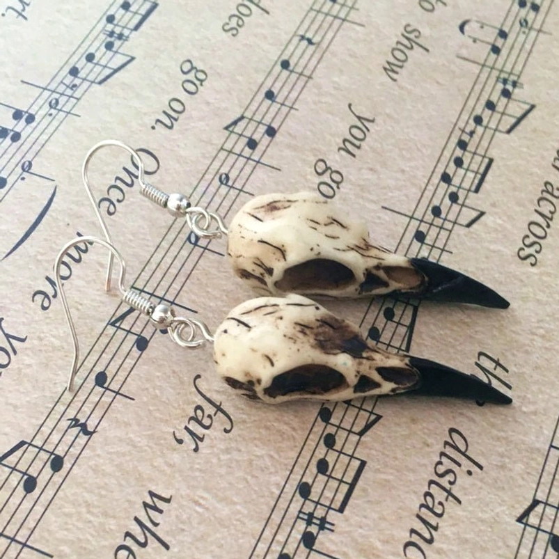 Flat lay overhead view of matching pair of gothic raven skull dangle earrings with dark brown weathered accents and silver wires on vintage music sheet