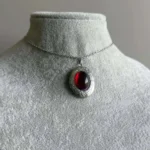Steampunk red cabochon pendant necklace displayed on jewelry mannequin neck, front view showing ornate engraved silver bezel with deep crimson stone against grey studio background