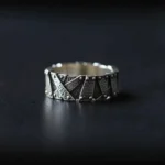 Industrial steampunk ring with riveted geometric patchwork design in silver-black metal, frontal flat lay on dark matte tabletop