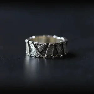 Industrial steampunk ring with riveted geometric patchwork design in silver-black metal, frontal flat lay on dark matte tabletop