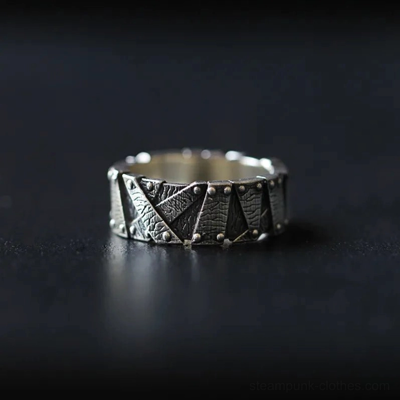 Industrial steampunk ring with riveted geometric patchwork design in silver-black metal, frontal flat lay on dark matte tabletop