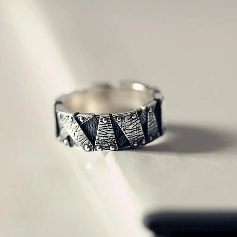 Three-quarter side perspective of industrial steampunk ring displaying silver-black riveted geometric pattern on light-colored desk