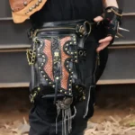 Steampunk Serpento black leather thigh holster bag with brass skull and crossbones hardware, rivets, and fish-scale texture worn on model's leg in industrial outdoor setting