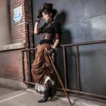 Victorian steampunk skirt with layered bustle design, brown suede with striped accents, worn with coordinating top hat and goggles by female model against industrial brick wall backdrop